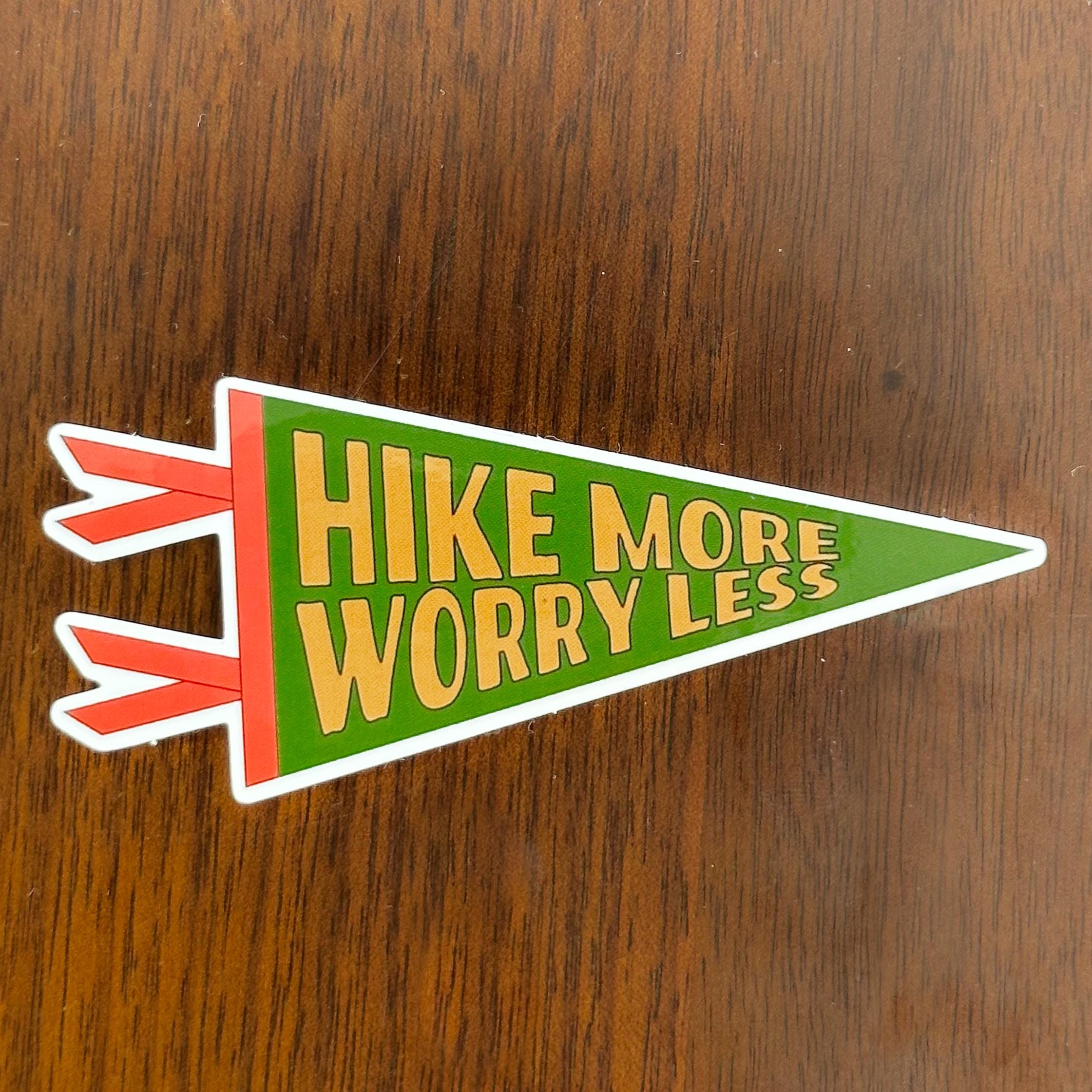 A green, gold and orange pennant-shaped sticker with 'Hike More Worry Less' text on a wooden surface