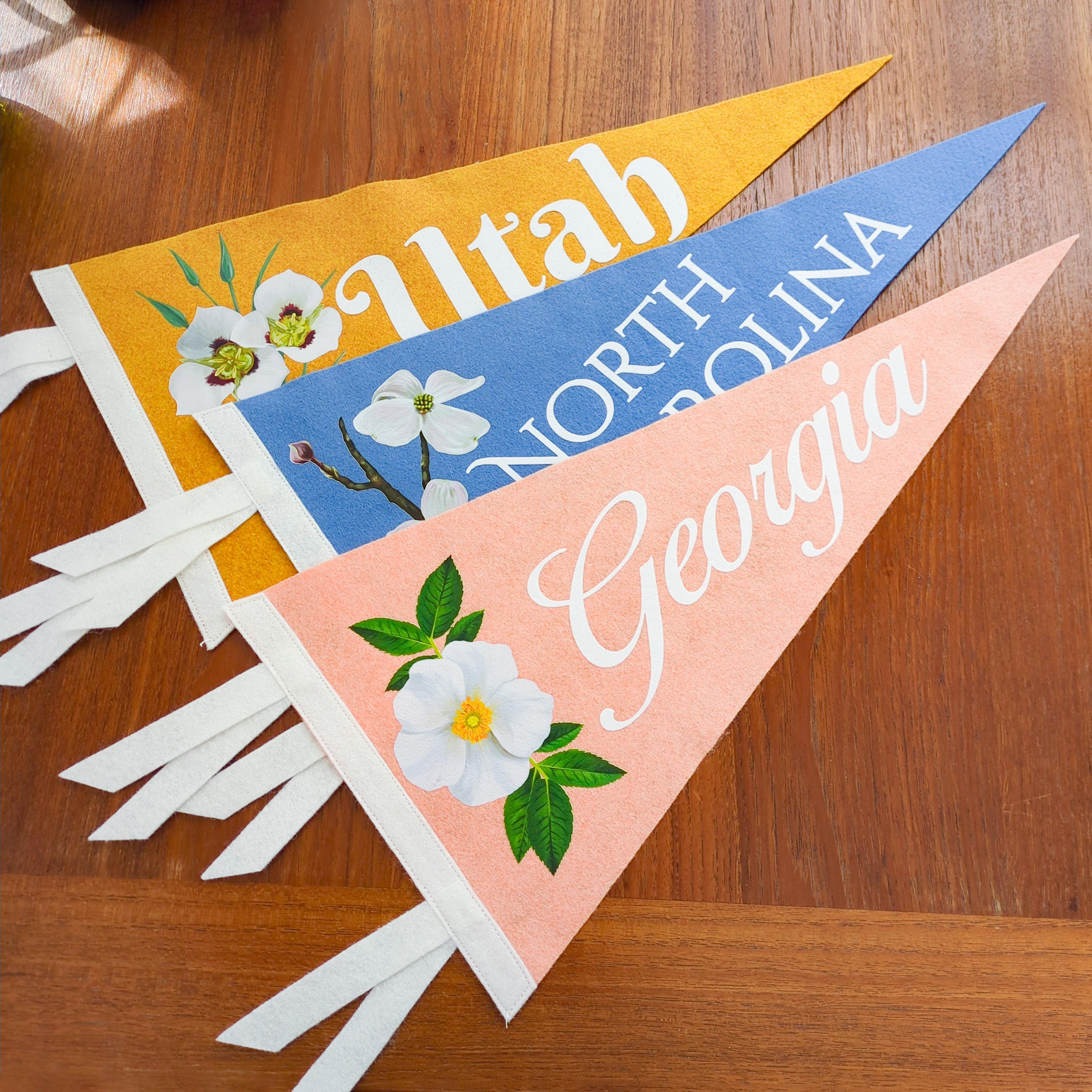 Three colorful pennant flags that say "Georgia", "North Carolina" and "Utah" with floral designs on a wooden surface