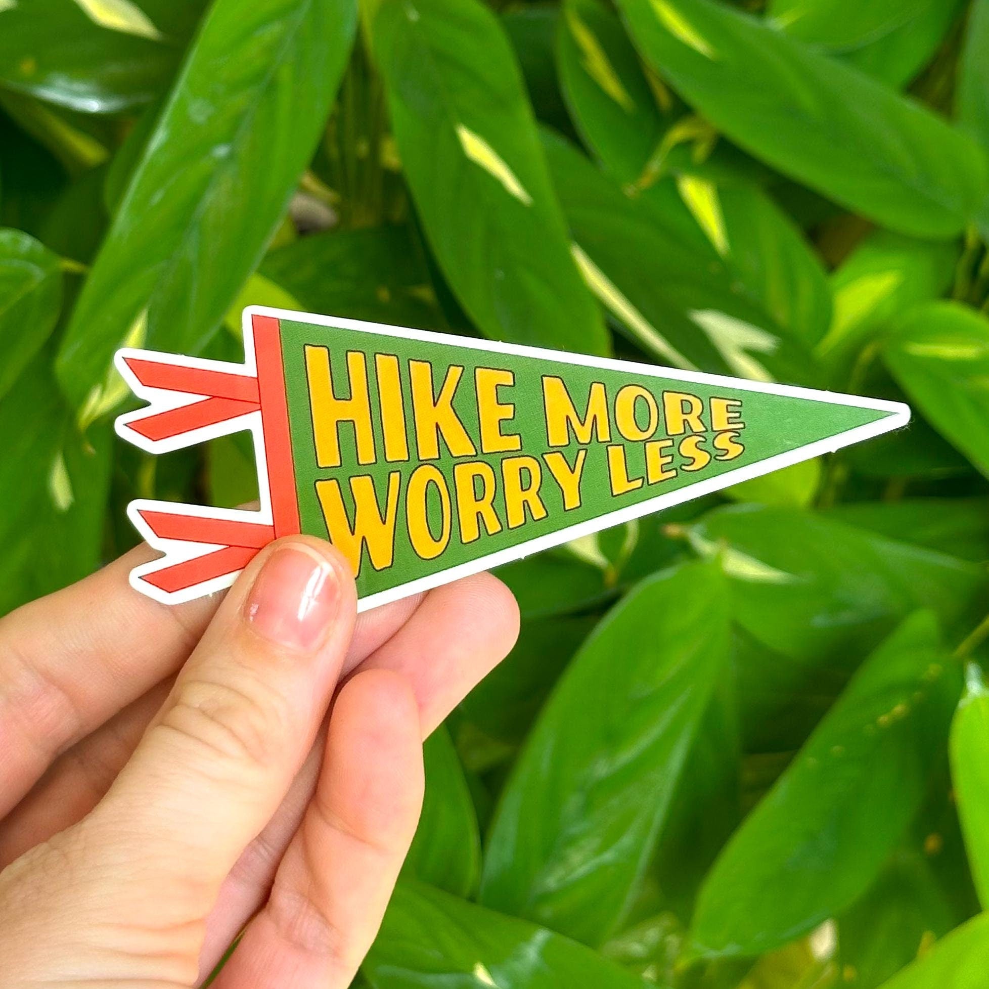 Hand holding a 'Hike More Worry Less'  pennant-shaped sticker against a green leafy background