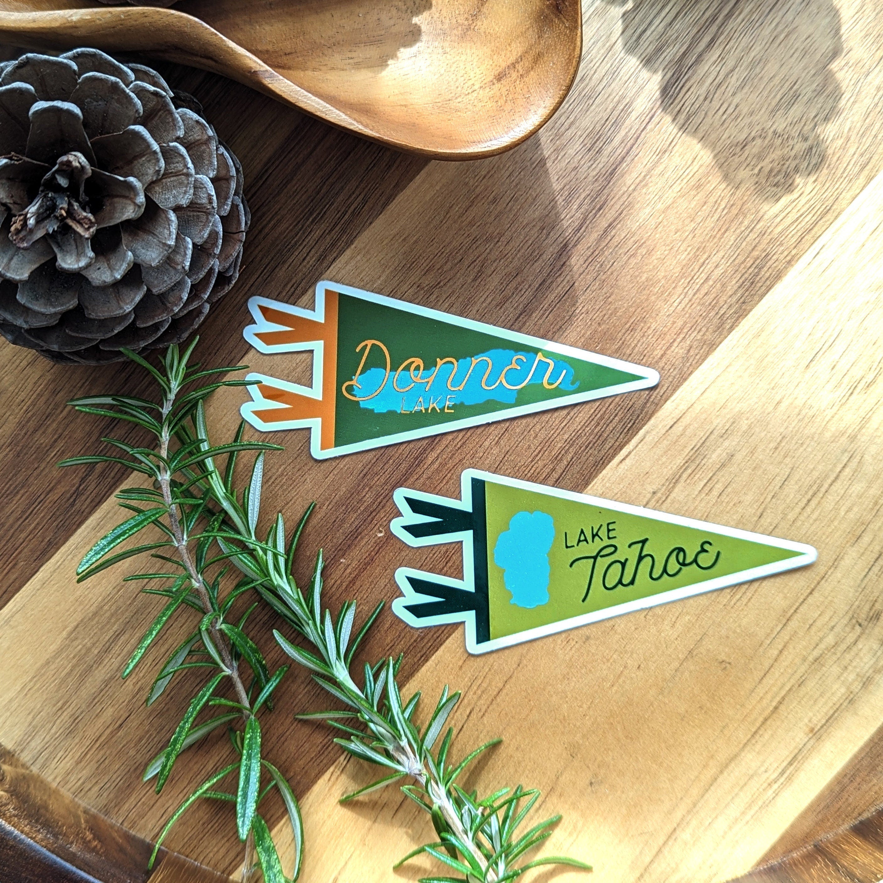 Two green Donner Lake and Lake Tahoe pennant shaped stickers on a wooden surface with a pine cone and rosemary.