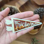 Hand holding a 'Truckee California' sticker. The sticker is shaped like a pennant with a rust red background, white text, a poppy flower design, and white trim.