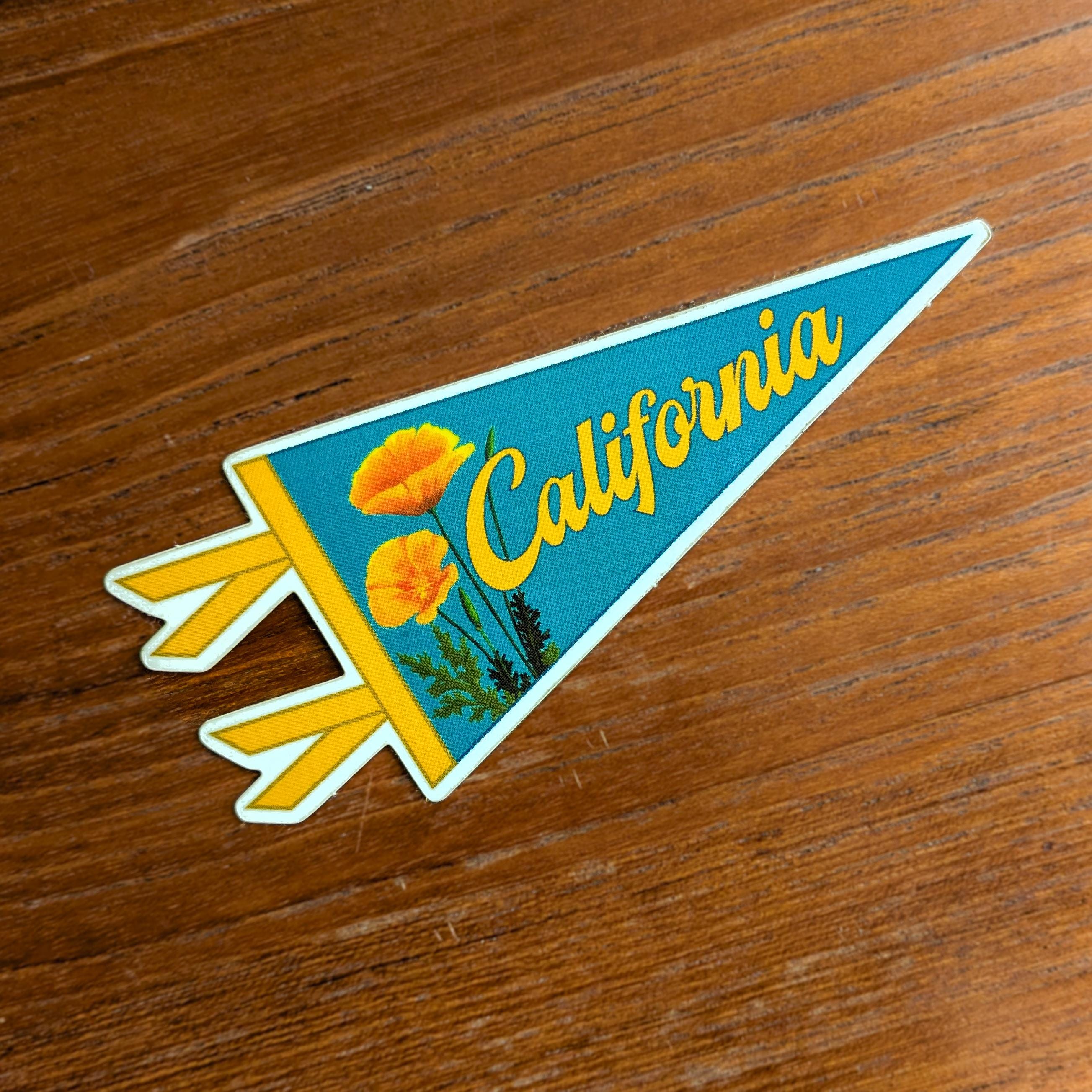 A blue pennant shaped sticker with 'California' text and a yellow and orange poppy design, displayed on a wood background.