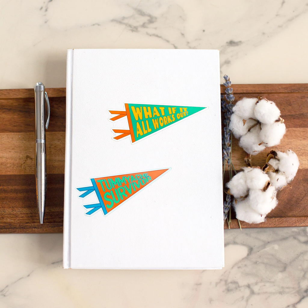 a noteboook with a colorful Pennant-shaped sticker that says "What if It All Works Out" , and a colorful pennant sticker that says "Tummyache Survivor"