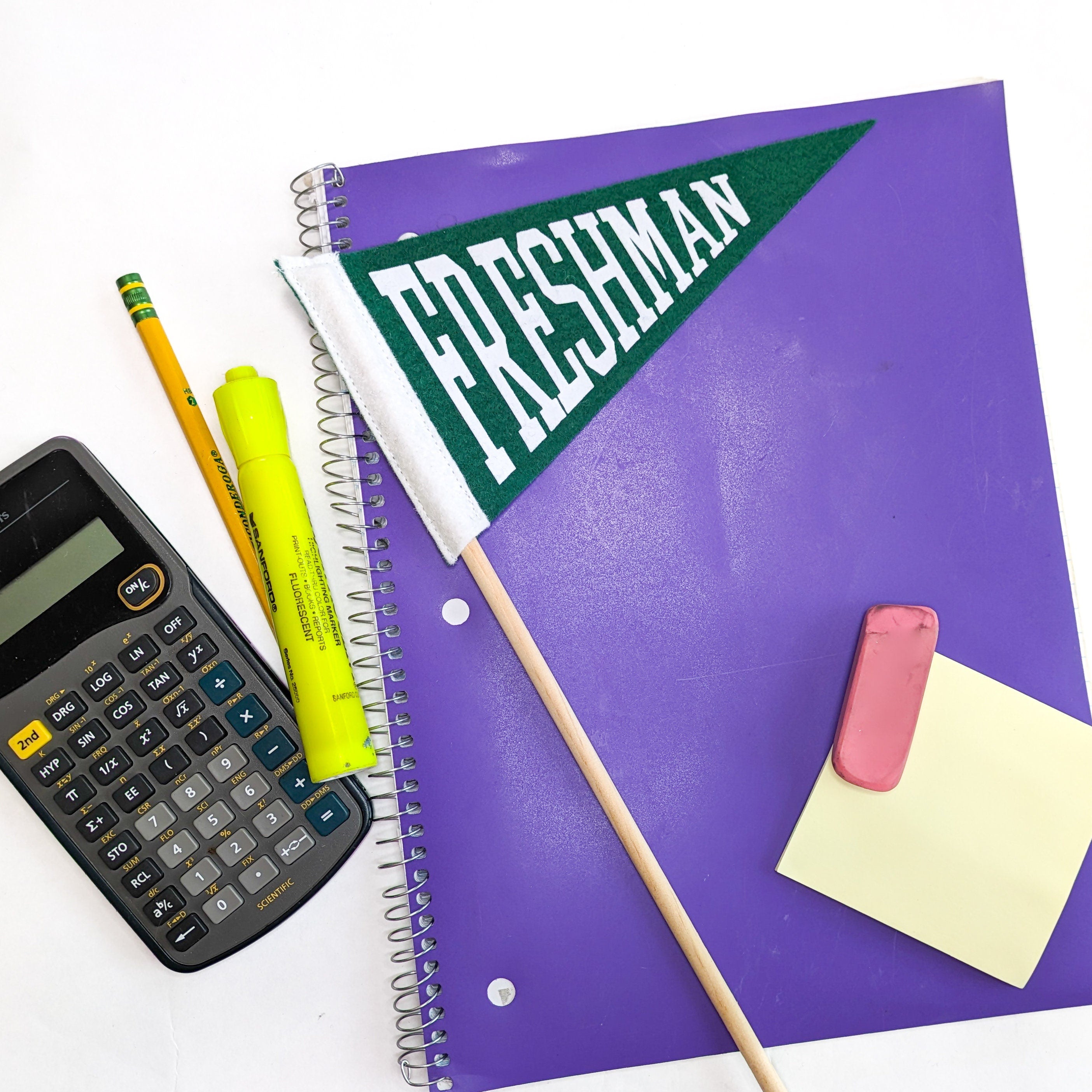 Purple notebook with a green 'Freshman' flag, calculator, markers, and other school supplies on a white background