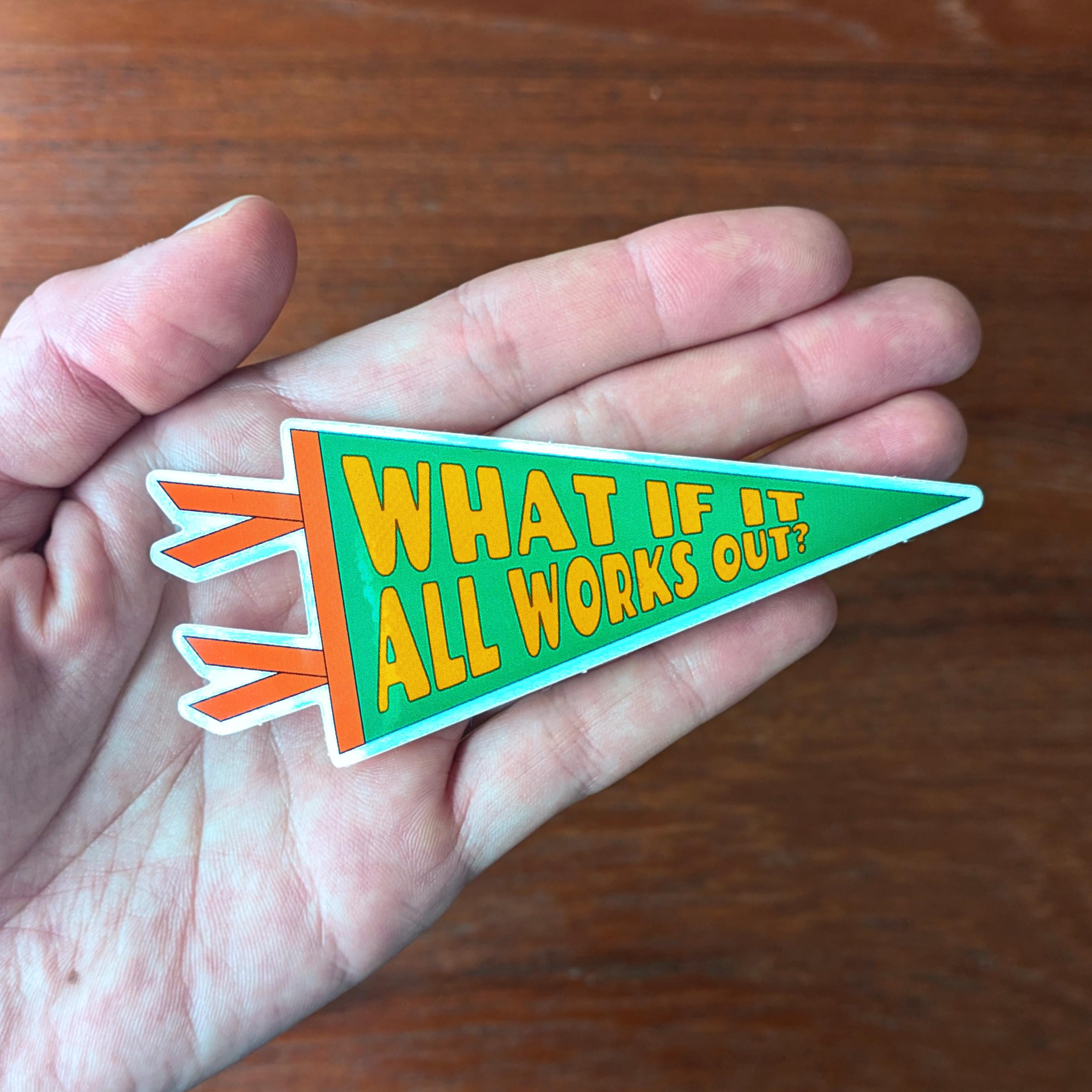 Hand holding a colorful Pennant-shaped sticker that says "What if It All Works Out" in gold bubble text on a green background, with orange trim.