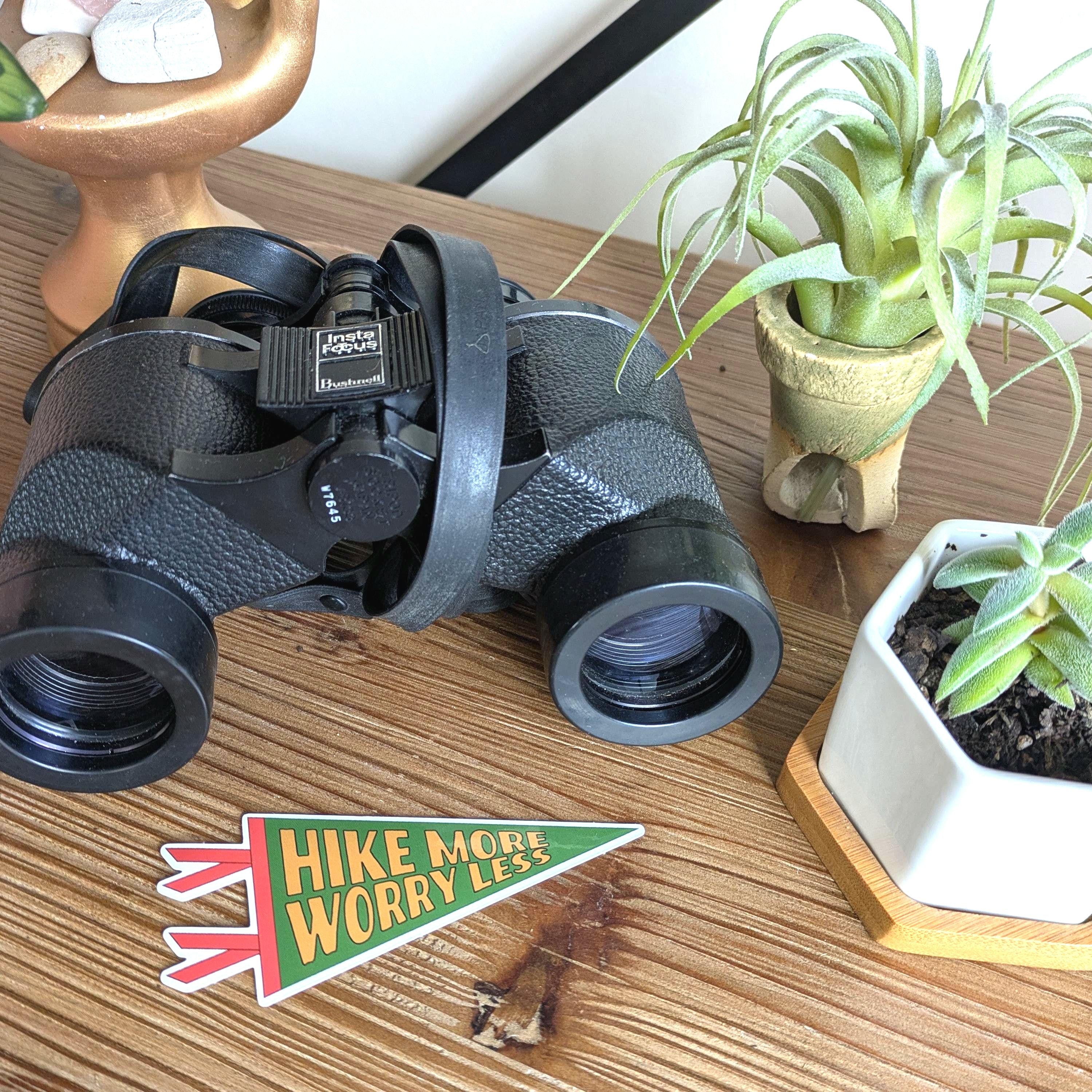 Binoculars and plants on a shelf with a 'Hike More Worry Less' sticker.