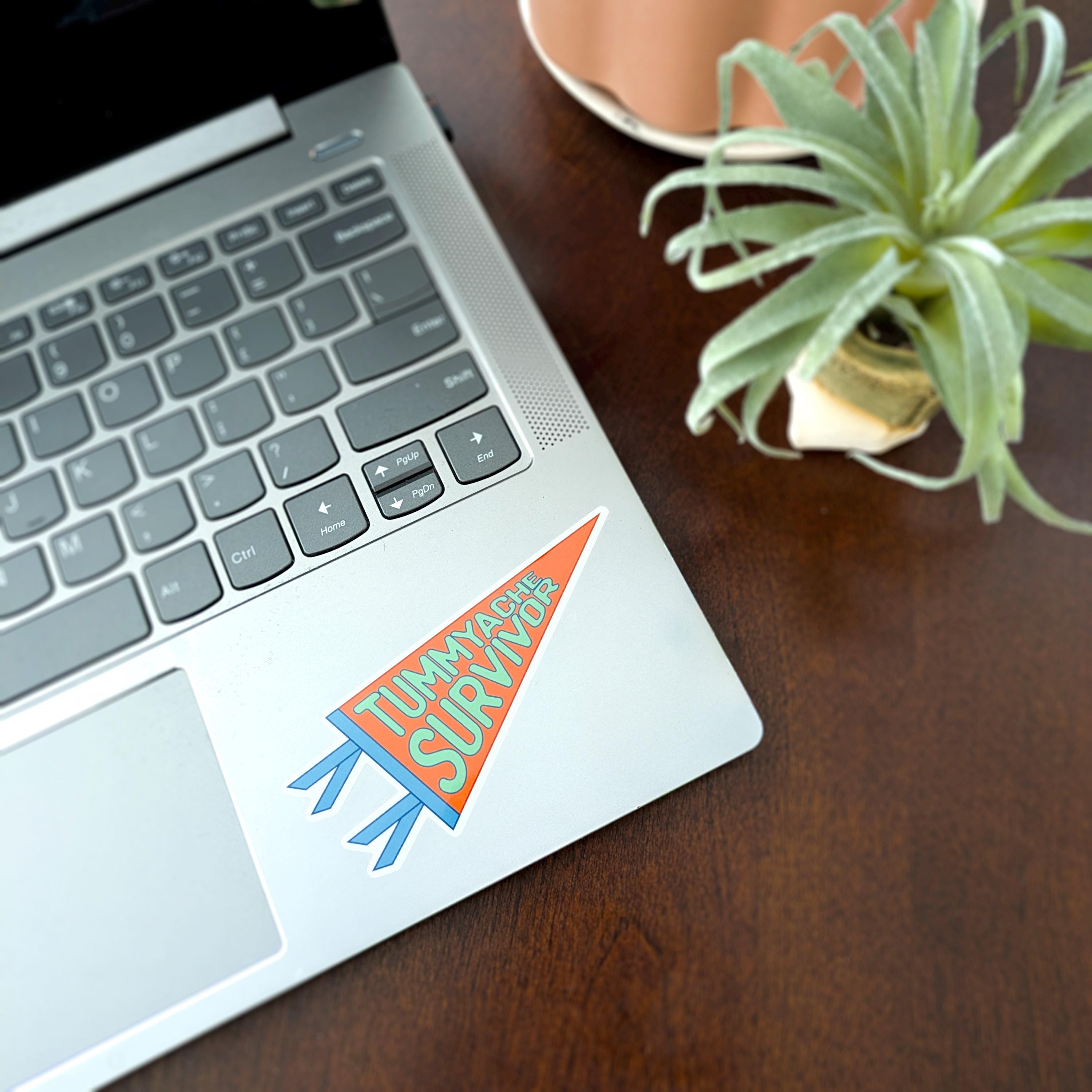 A laptop with a pennant-shaped Tummyache Survivor sticker on it.