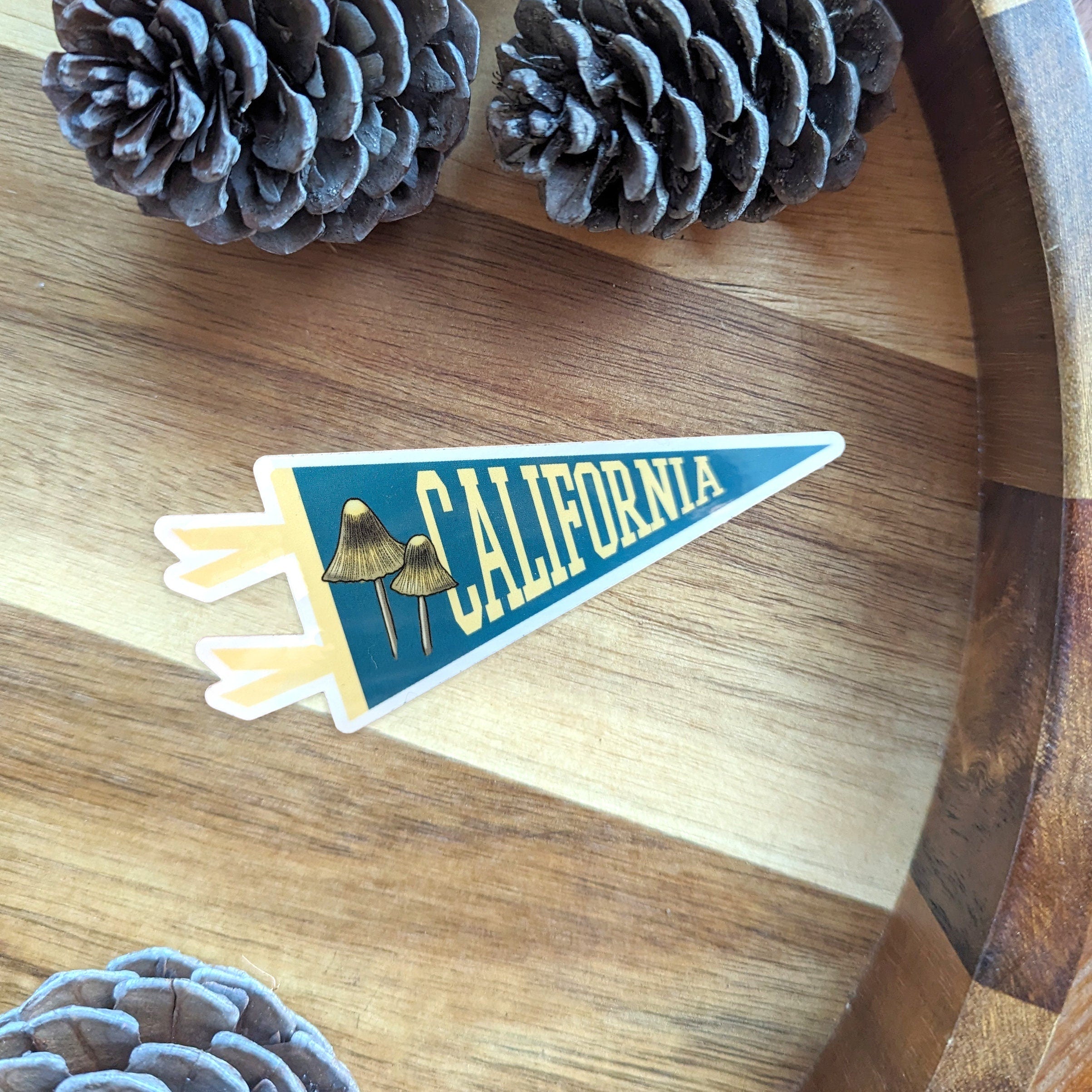 Pennant-shaped blue and yellow sticker with 'California' text and a mushroom illustration displayed on a wooden surface 