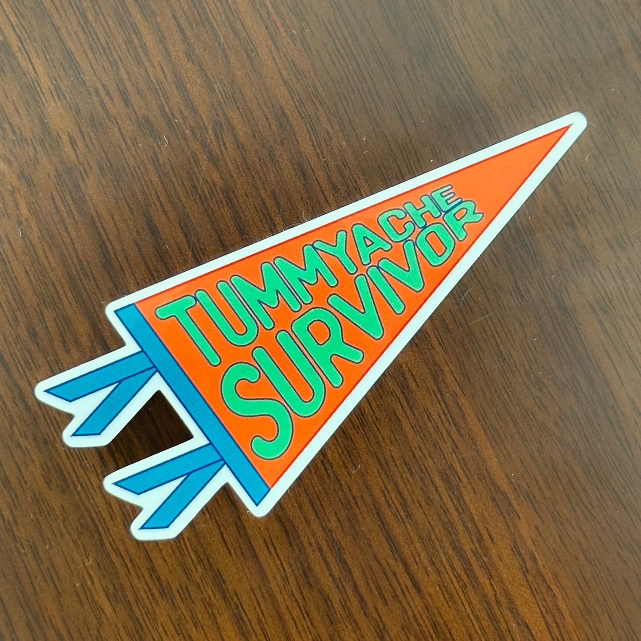 A pennant shaped sticker that says "Tummyache Survivor" in bright green bubble text on an orange background, with blue trim.