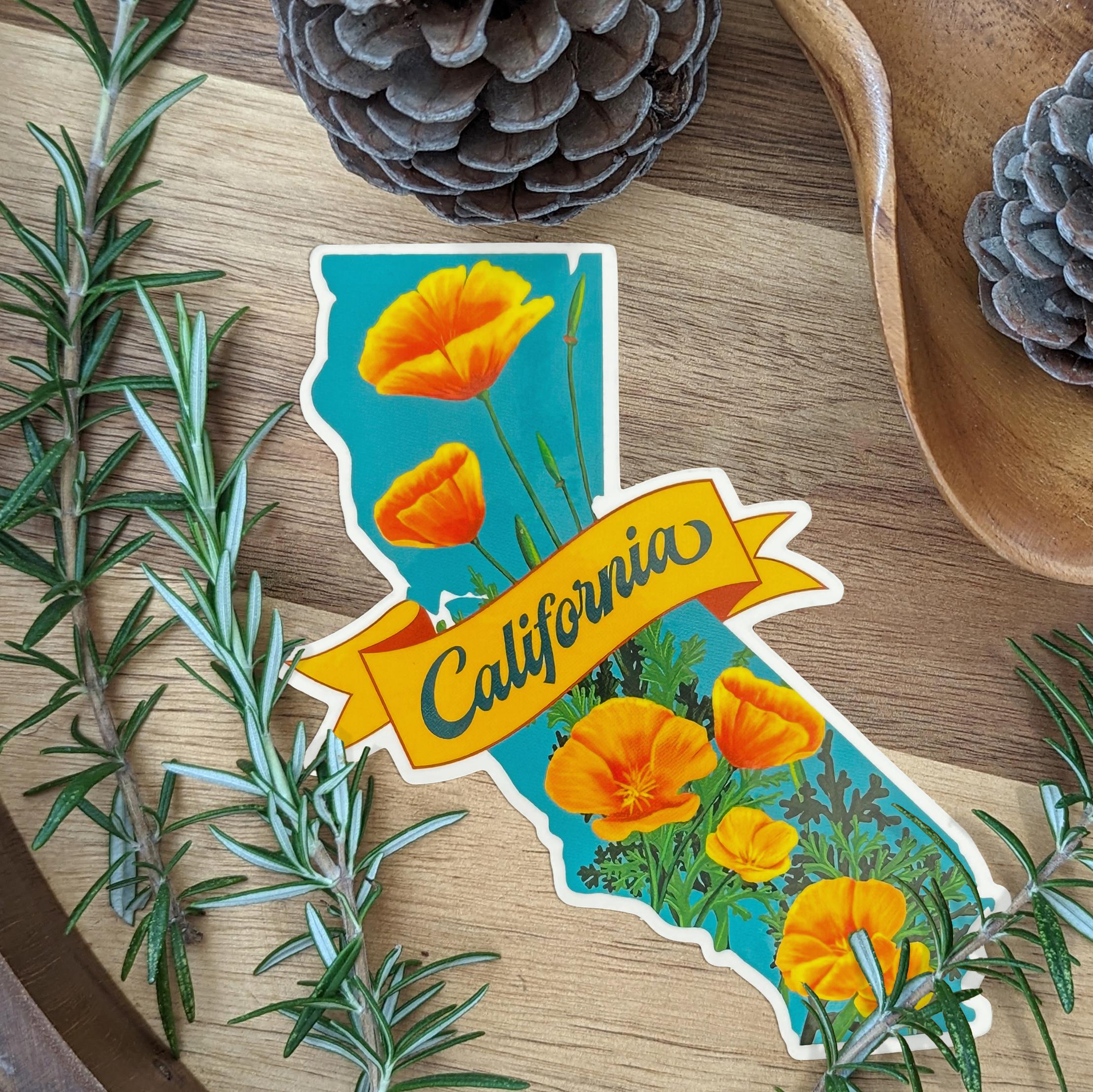 Sticker in the shape of  California with poppies on it, displayed on a wooden surface with plants