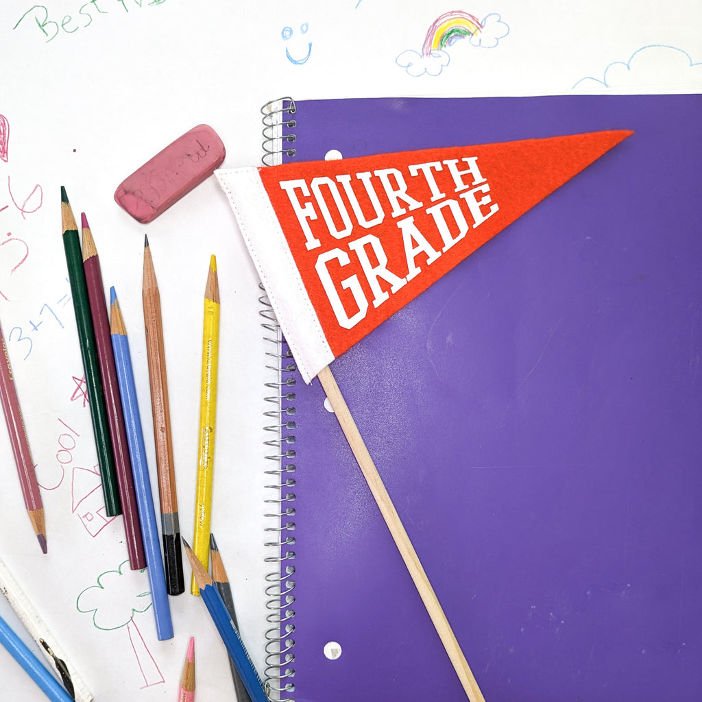Purple notebook with a 'Fourth Grade' pennant, surrounded by colorful pencils and an eraser on a white background with drawings.