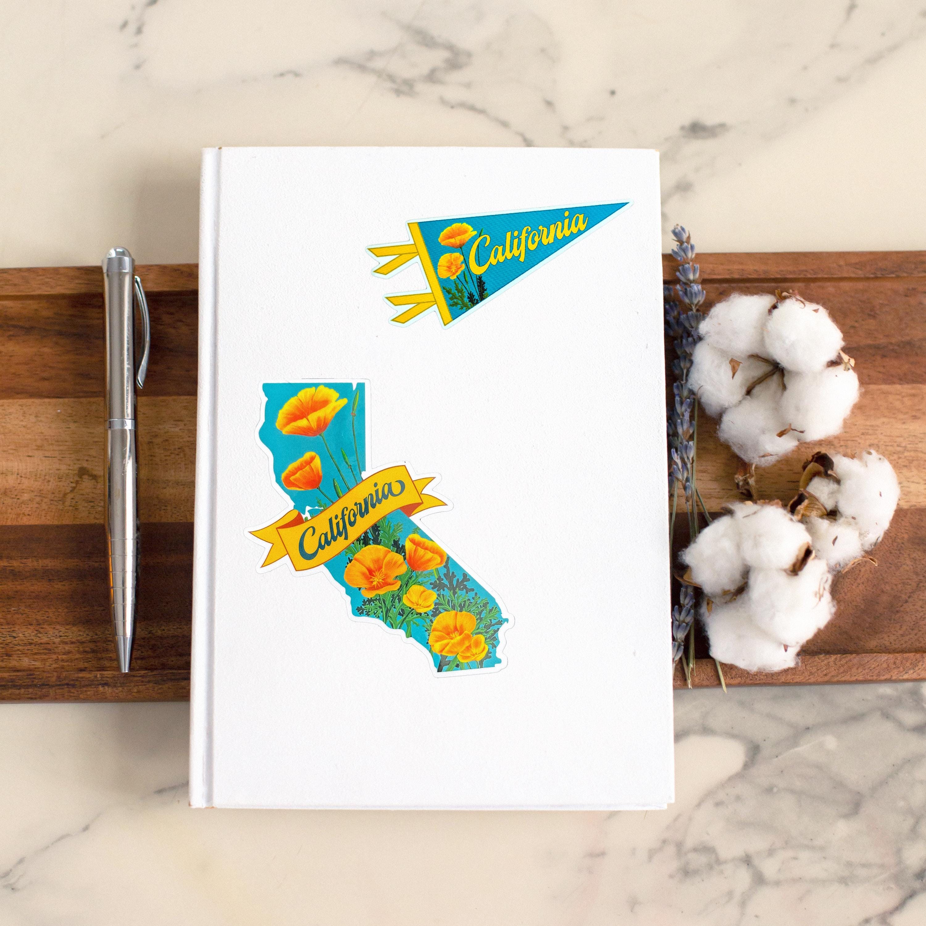 A notebook with two sticker on it. A smaller pennant-shaped blue and orange sticker that says "California" with a poppy flower design, and a larger California-shaped sticker.