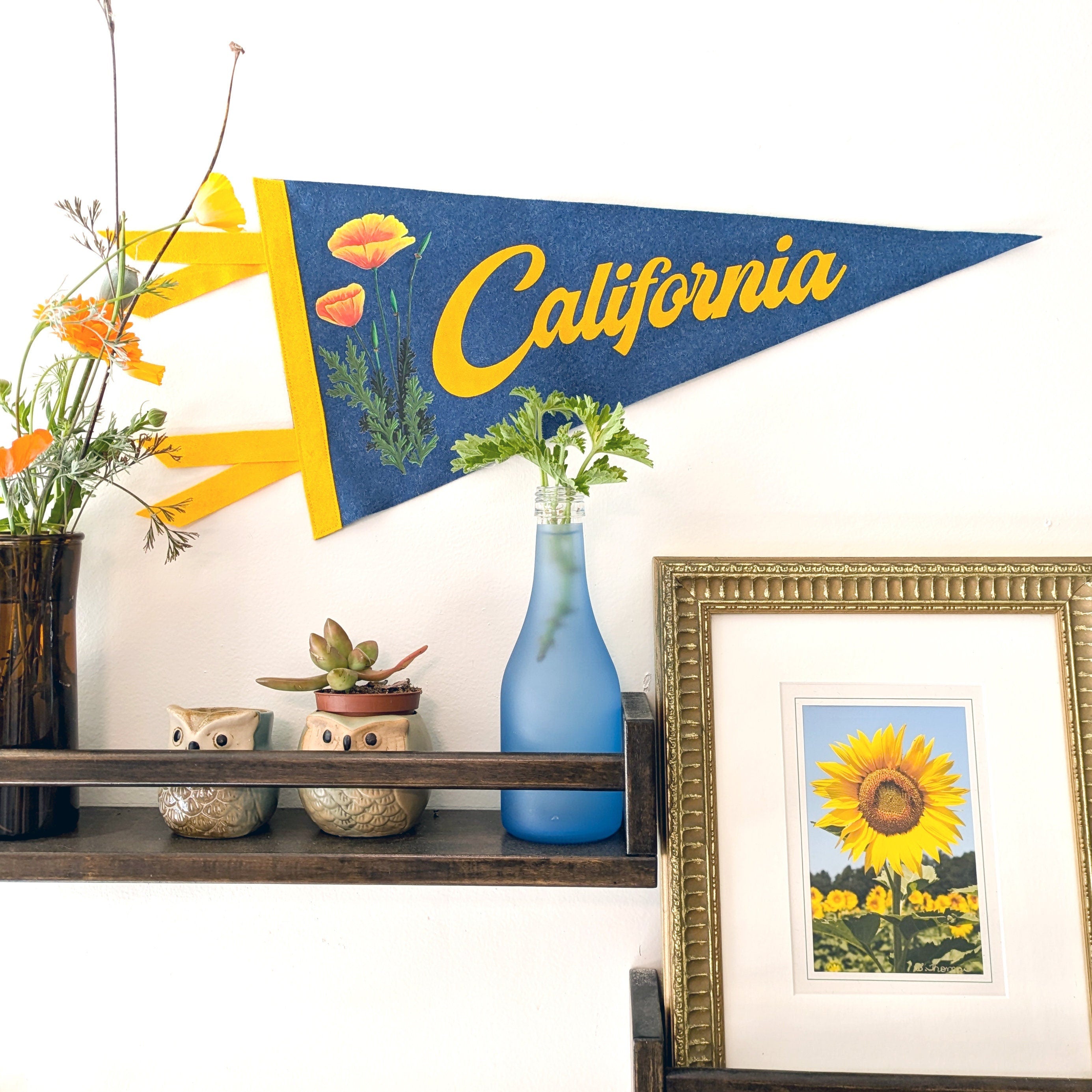 Decorative felt pennant with 'California' text in yellow on a blue background, placed above a shelf with decorative items.