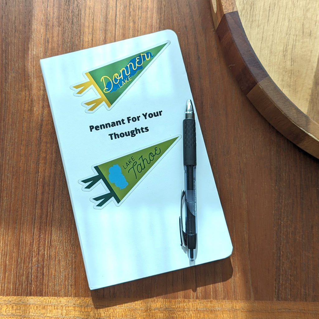 Notebook with pen on a wooden surface, with a donner lake and lake tahoe sticker on it.