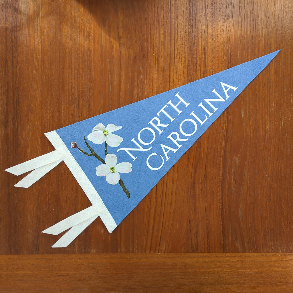 North Carolina-themed pennant with white dogwood flowers and white text on a light blue felt background