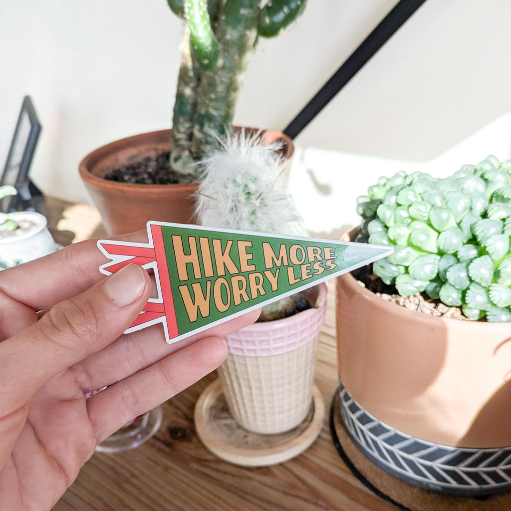 Hand holding a pennant-shaped sticker with 'Hike More Worry Less' text in front of potted plants.