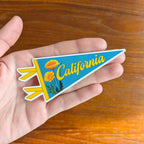 Hand holding a 'California' pennant-shaped blue and orange sticker with flowers 