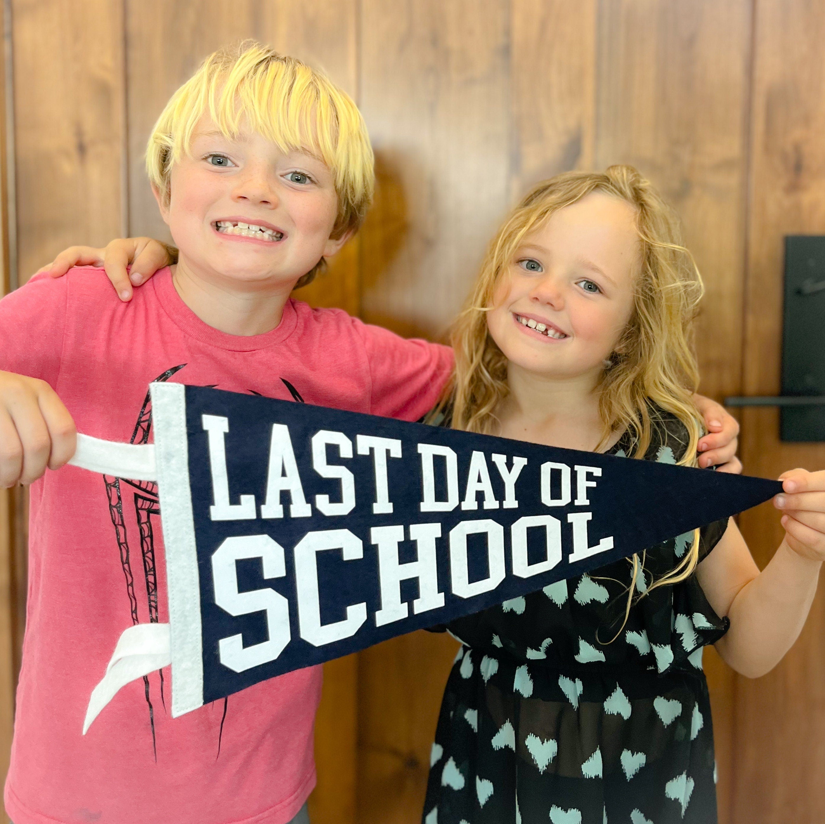 Two children holding a 'Last Day of School' sign in front of a wooden wall.