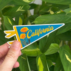 Hand holding a ''California' pennant-shaped blue and orange sticker with flowers  against a green leafy background