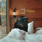 Person reading a book in bed with a wooden wall and window view of trees in the background, with a "Utah" flag on the wall