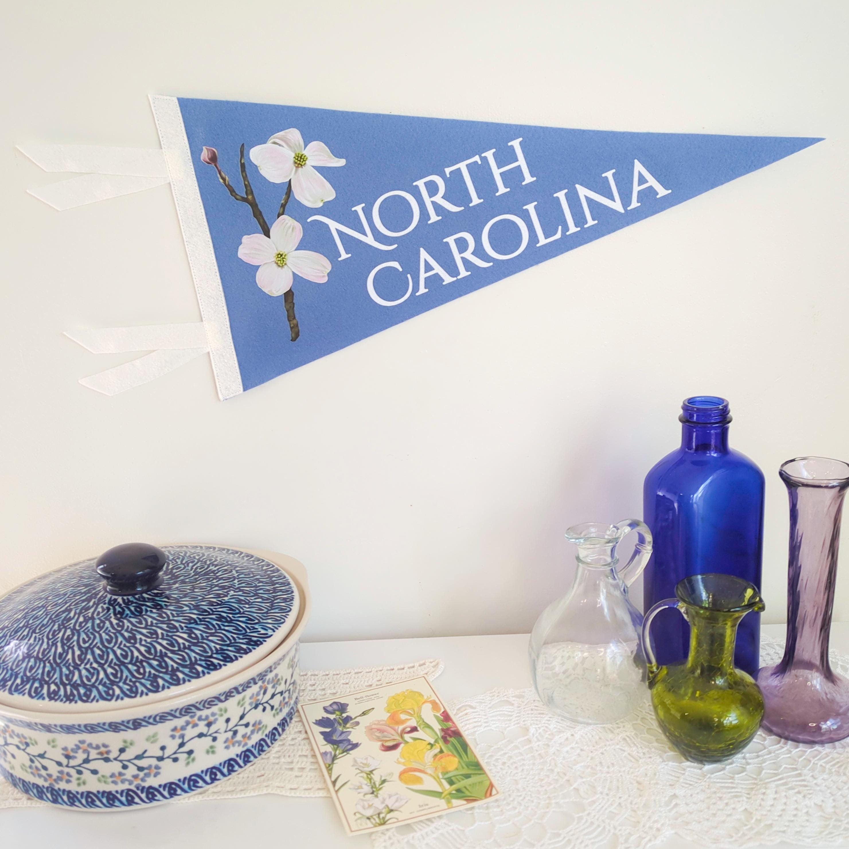 Decorative setting with a North Carolina flag, blue ceramic dish, glass bottles, and a card on a white surface.
