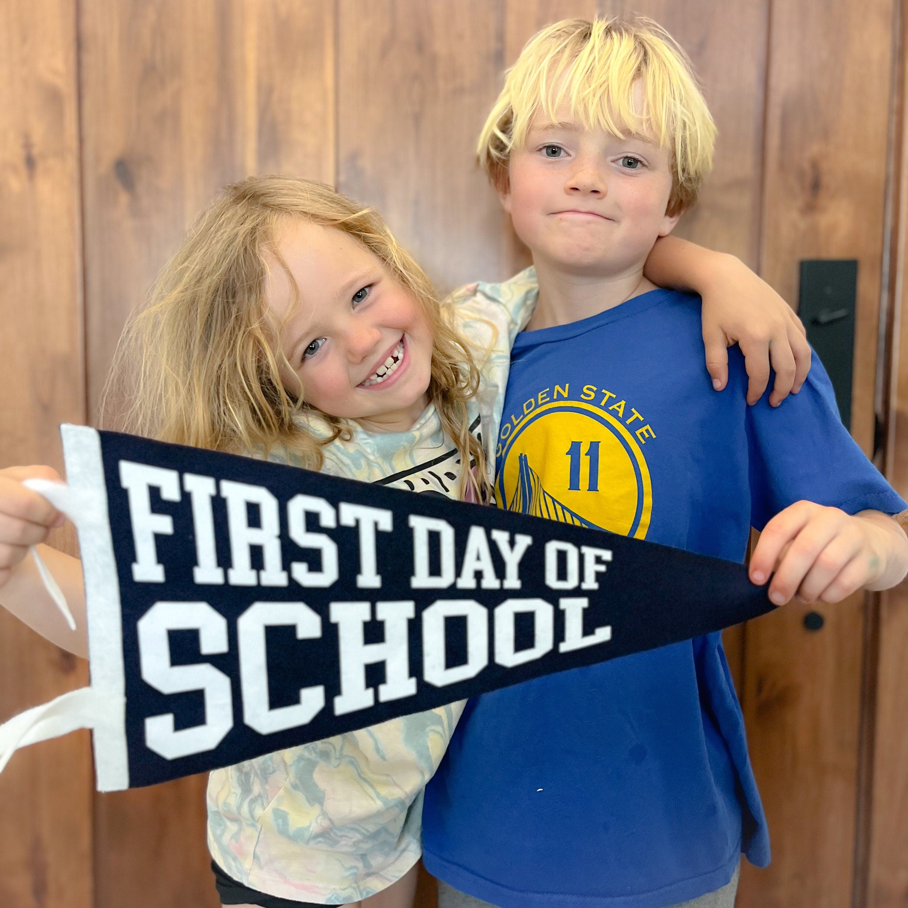 Two children holding a 'First Day of School' sign in front of a wooden wall.