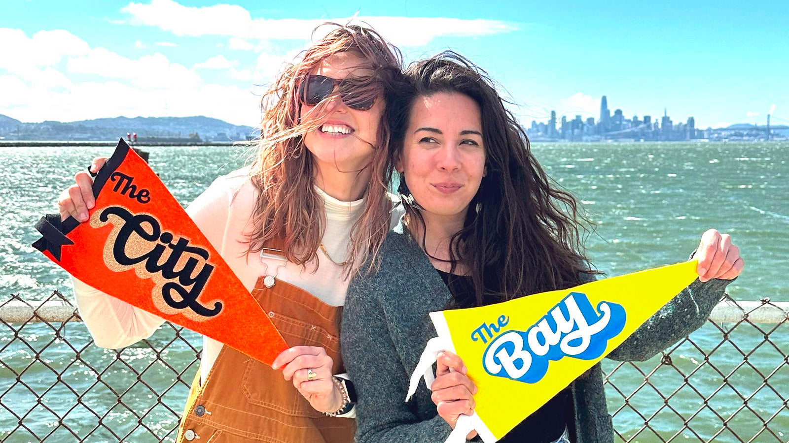 Two women holding 'The City' and 'The Bay' pennant flags with a scenic background of water and skyline.